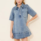 A woman modeling a light blue denim shirt dress with short sleeves, a buttoned collar, and a chest pocket, paired with black Mary Jane flats, against a plain beige studio background.